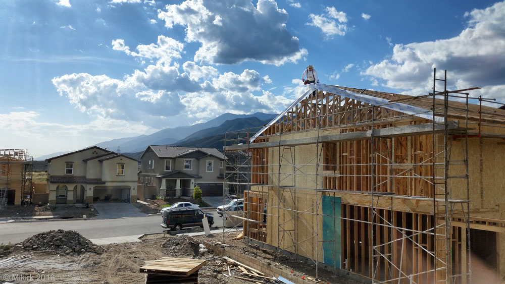 A construction worker stands on the roof of a wood frame house on a sunny day. The building doesn't have walls yet, so we see into the structure of the home, with scaffolding lining the outside. In the background, we see two suburban homes, and a third also under construction.