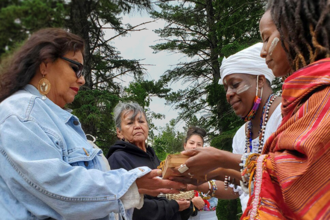 Two women, a Black woman and an Indigenous woman, trade ceremonial tokens as others watch. They stand amongst tall pine trees.