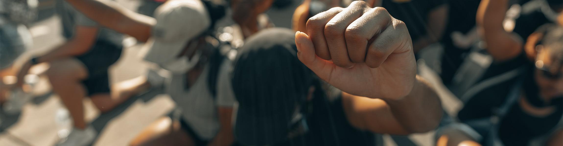 A closeup photograph of a Black protestor raising their fist, surrounded by many other protestors