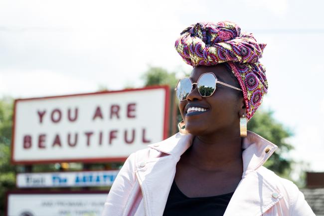 A young Black woman with a purple-yellow paisley hair wrap and big sunglasses smiles. A blurry sign behind her reads "YOU ARE BEAUTIFUL."