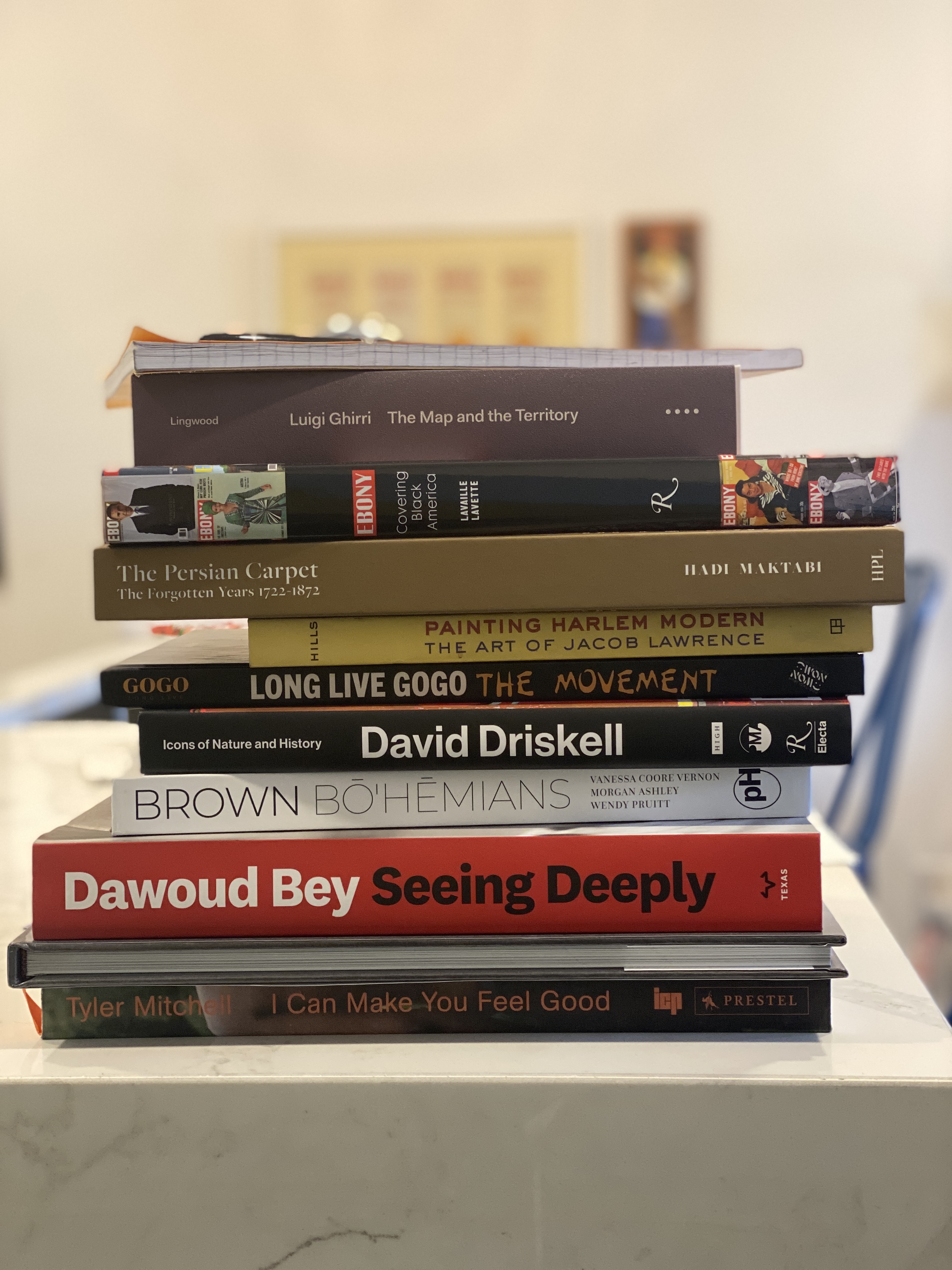 A stack of 11 books and notebooks with the bindings in view. Most are about art, culture, and representation. Among the books are Dawoud Bey's 'Seeing Deeply,' Patricia Hill's 'Painting Harlem Modern,' and James Lingwood's book on Luigi Ghirri 'The Map and the Territory.'