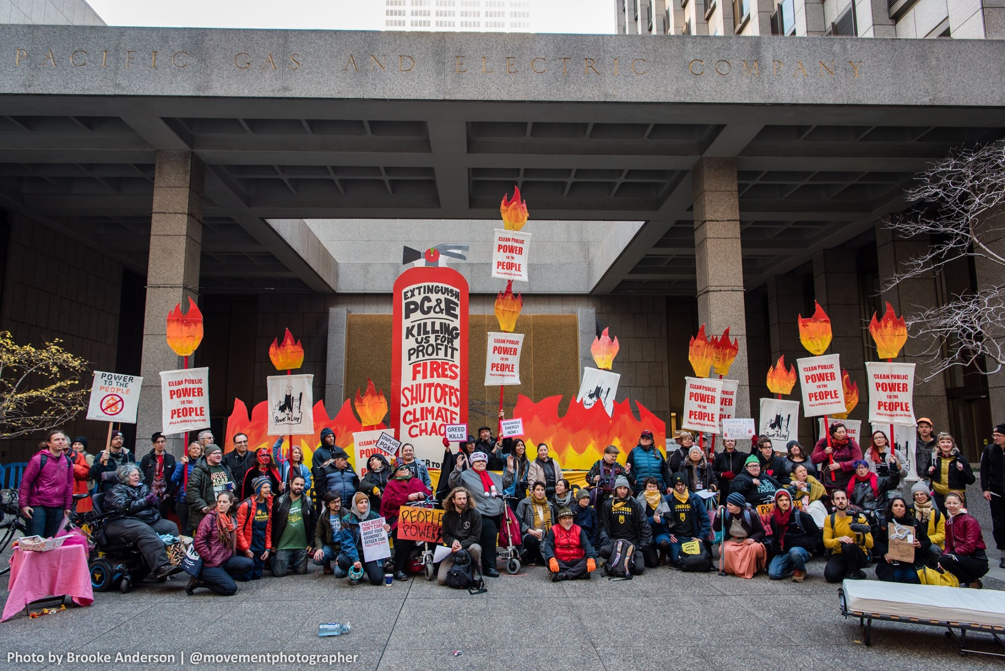 A large group of about 80 climate justice protestors in front of PG&E headquarters. The protestors hold banners and placards reading Power to the People. A large cardboard fire stands behind them, and an extinguisher reads Extinguish PG&E Killing us for Profit. Many of the protestors present are in wheelchairs or use mobility aids.
