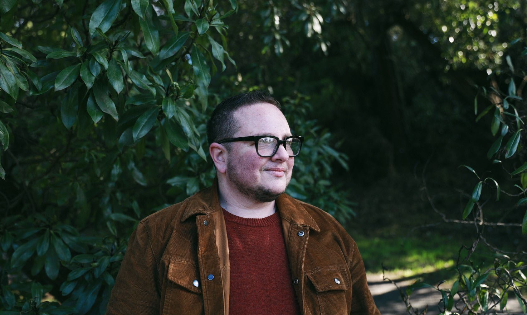 Photograph of Elliot Kukla, a white Jewish man with short slicked hair, black thick-rim reading glasses, and a corduroy jacket. He gazes somewhere off camera; behind him is lush and green.