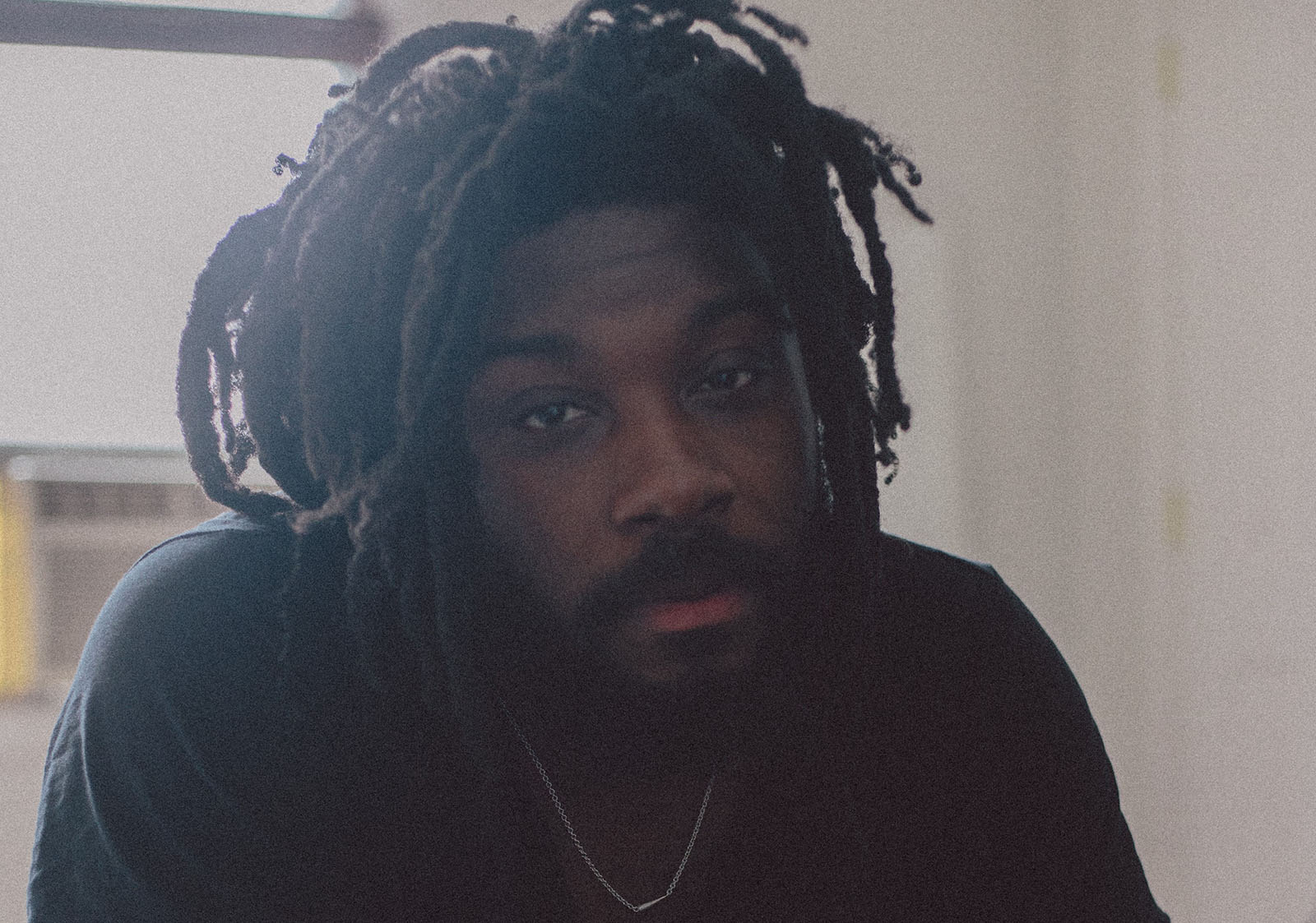 Photograph of Jason Reynolds, a Black man with dreadlocks and a full beard, wearing a black t-shirt. He gazes dreamily into the camera as soft white light peers out behind him.