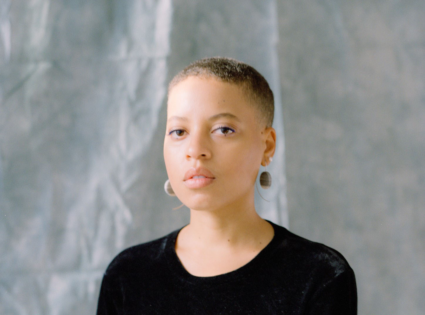 Photograph of Naima Green, a Black woman with short cropped hair, gazing softly into — almost through — the camera. She wears a black velour top, big stone earrings, and light eye makeup.