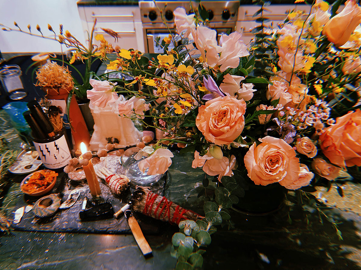 a still life image from Naima; a bursting bouquet of roses and other flowers occupies much of the frame. a lit candle, half-burnt sage, and other items cover the table top too.