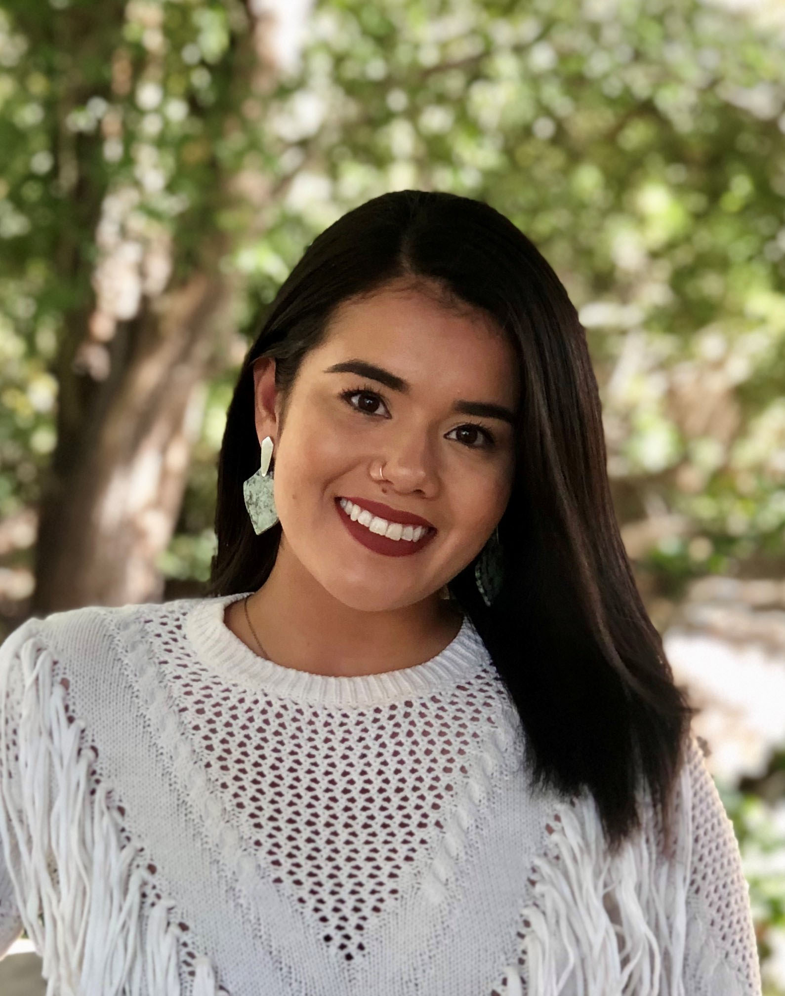 Photograph of Perla Torres, A portrait of Perla Torres, a young Latina with shoulder-length, smooth dark brown hair. She wears dark red lipstick, a knit top, a nose ring, and big stone earrings as she sweetly smiles a big grin towards the camera.
