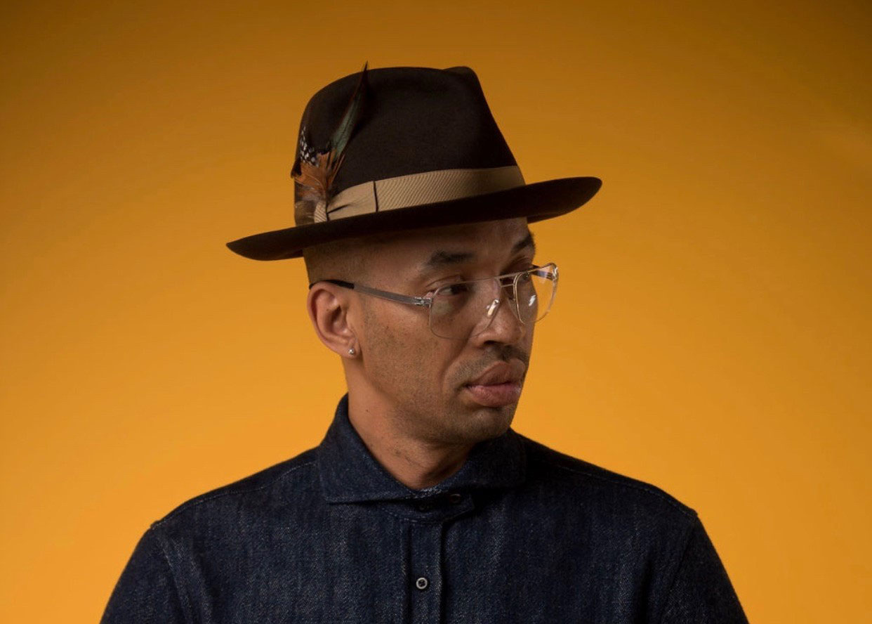 Photograph of Rich Medina, a Black man wearing a homburg hat with a feather, hip reading glasses, and a button-up shirt. He stands on a yellow-orange backdrop with his head cocked to the side, looking off camera.