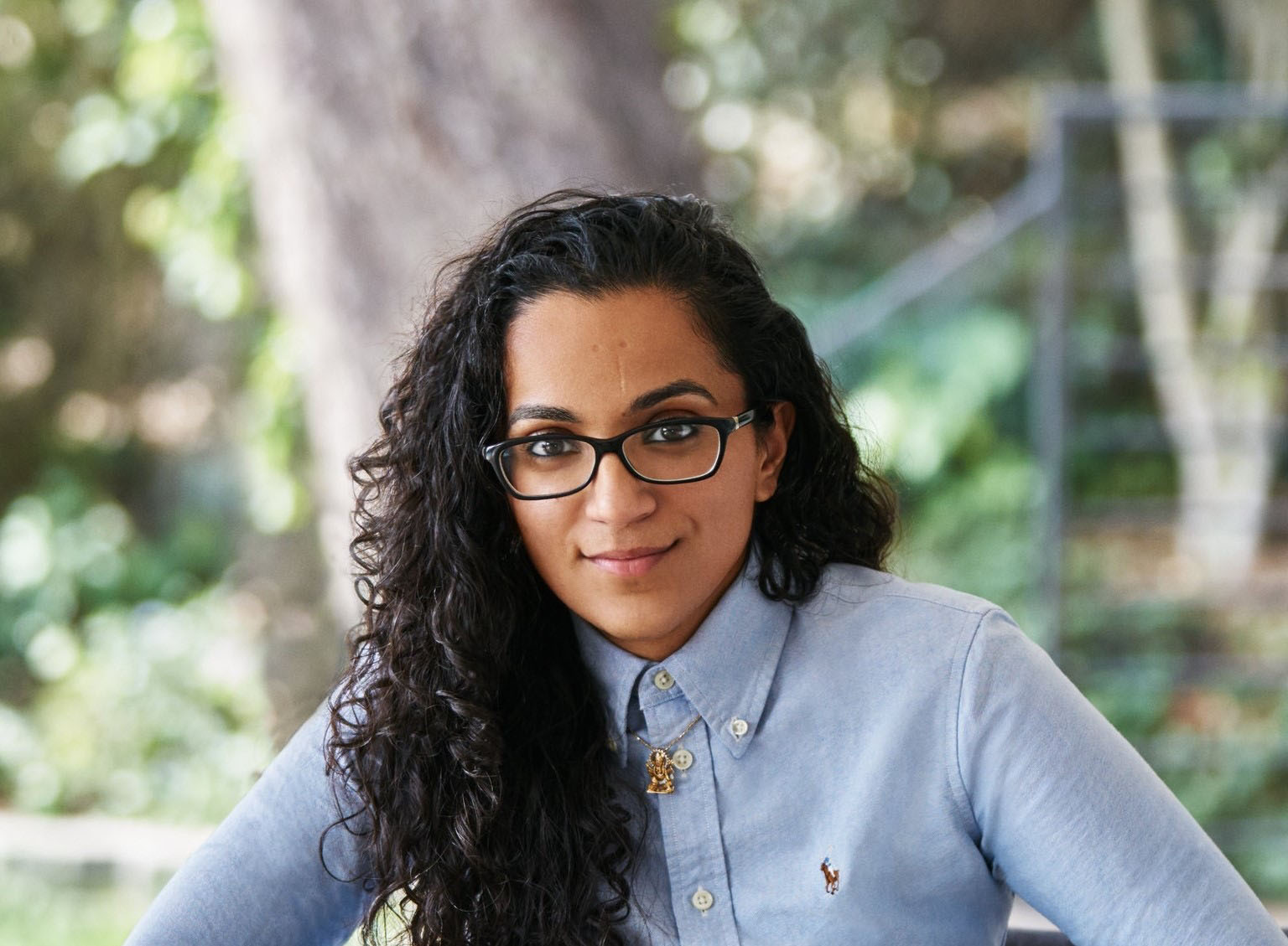 Photograph of Sonya Passi, a Brown woman with long wavy black hair. She wears a light blue buttonup shirt with a chain hanging under the color. She leans forward and smiles, arms on a table and fingers interlaced.