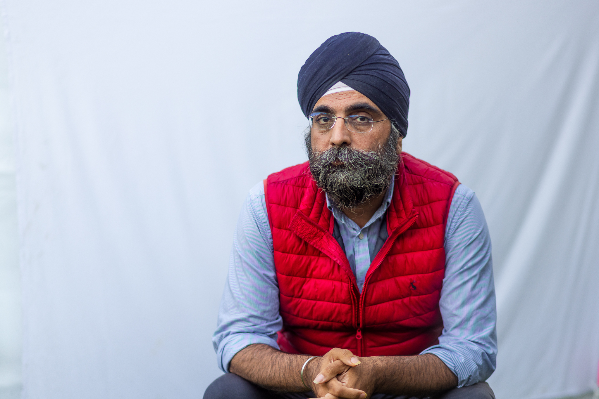 A Sikh man with a blue turban, red puffy vest, and blue button up shirt sits with hands clasped.