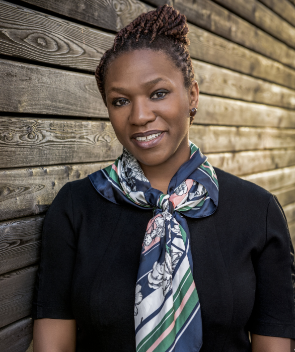 A Black woman with braided, tied-up hair and a patterned scarf stands smiling