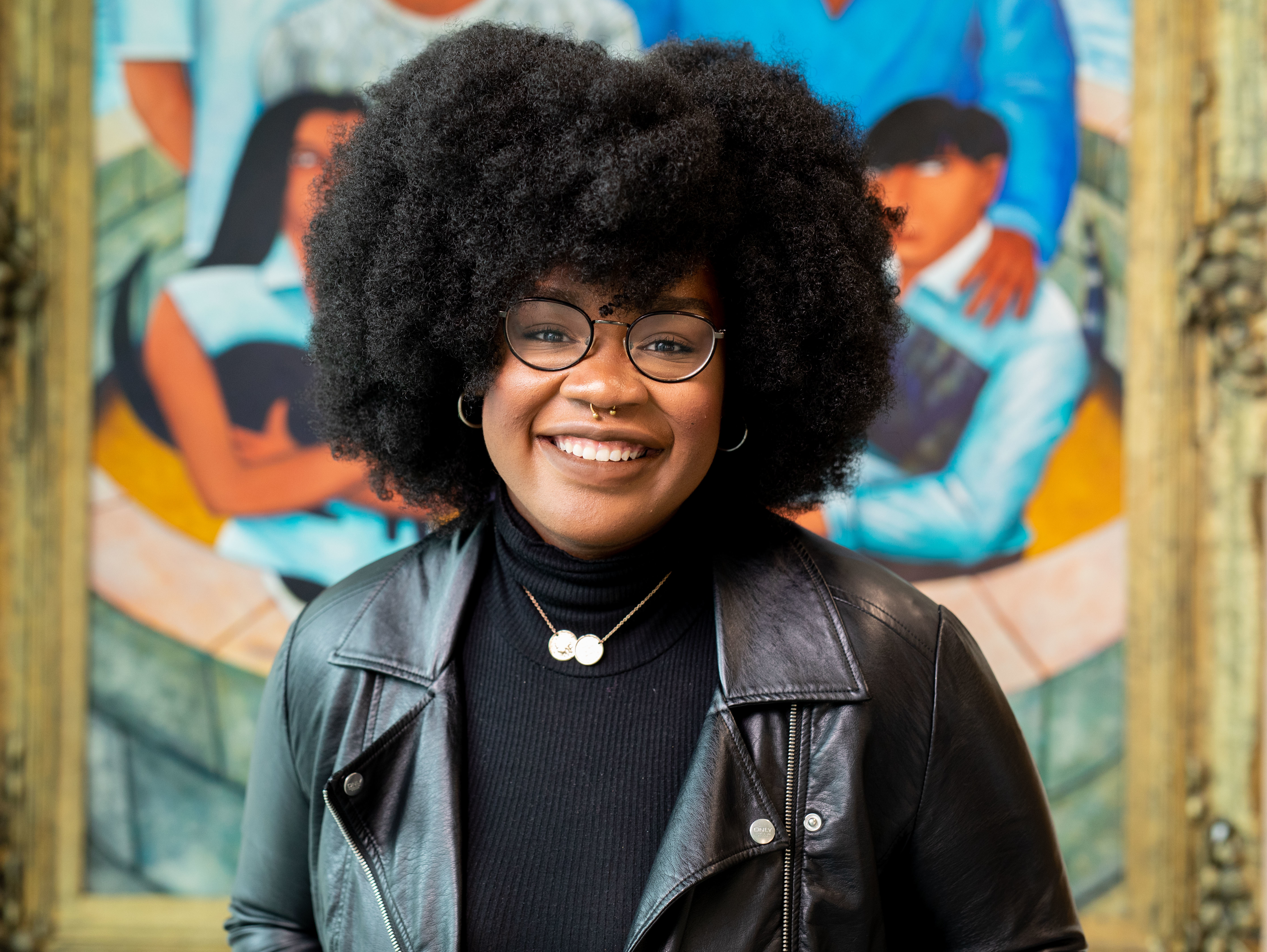 A Black woman with big dark hair and a leather jacket stands smiling in front of a mural.
