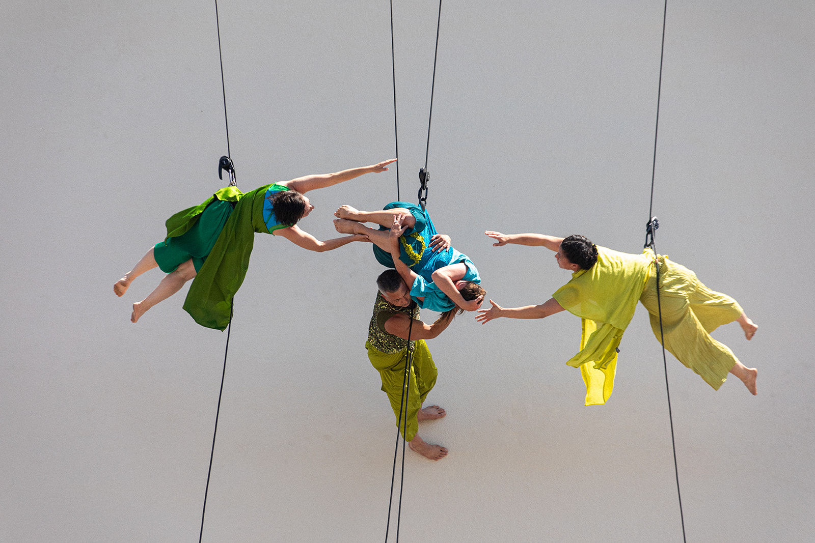 A group of dancers wearing jade green and teal blue dance while rappeling down a building