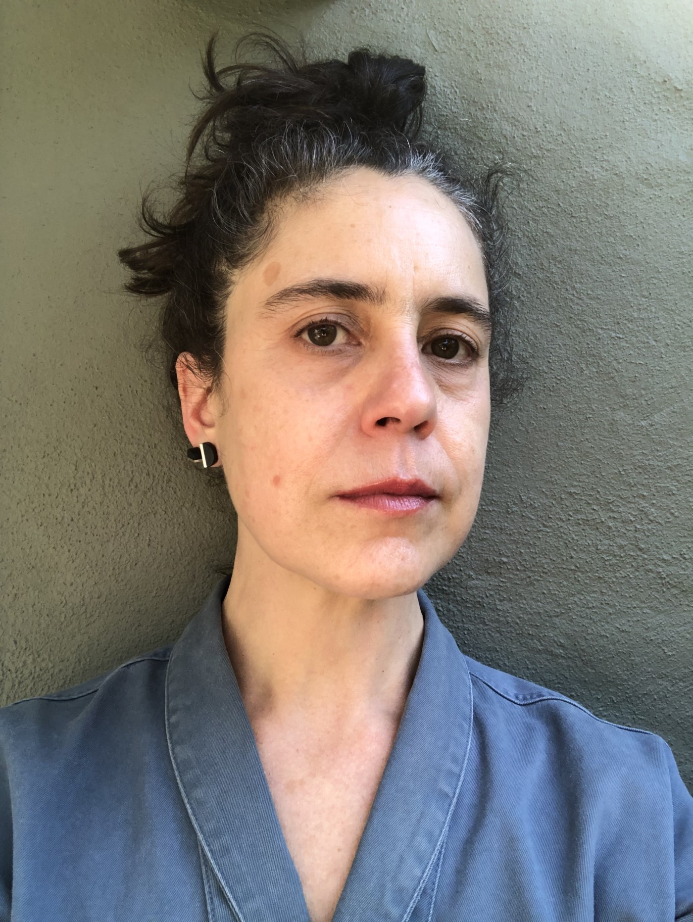 Natalia Brizuela is a woman with light skin, long pinned up black hair with white streaks, and a gentle demeanor. She wears a blue-grey blouse.