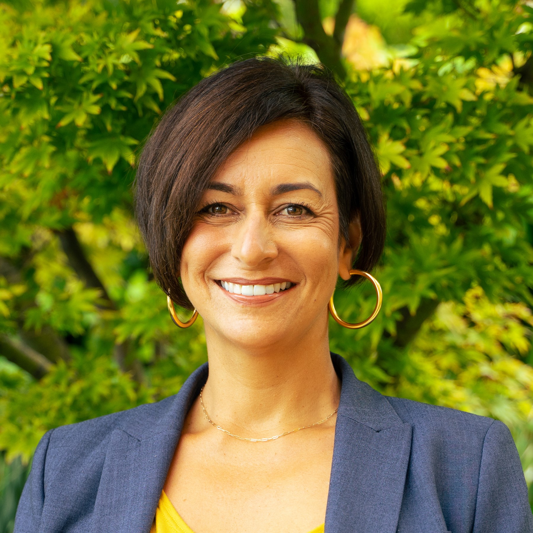 Kristina's headshot, she is a Latina woman with a short angled bob haircut and a warm smile. She wears gold hoop earrings and a blue suit jacket