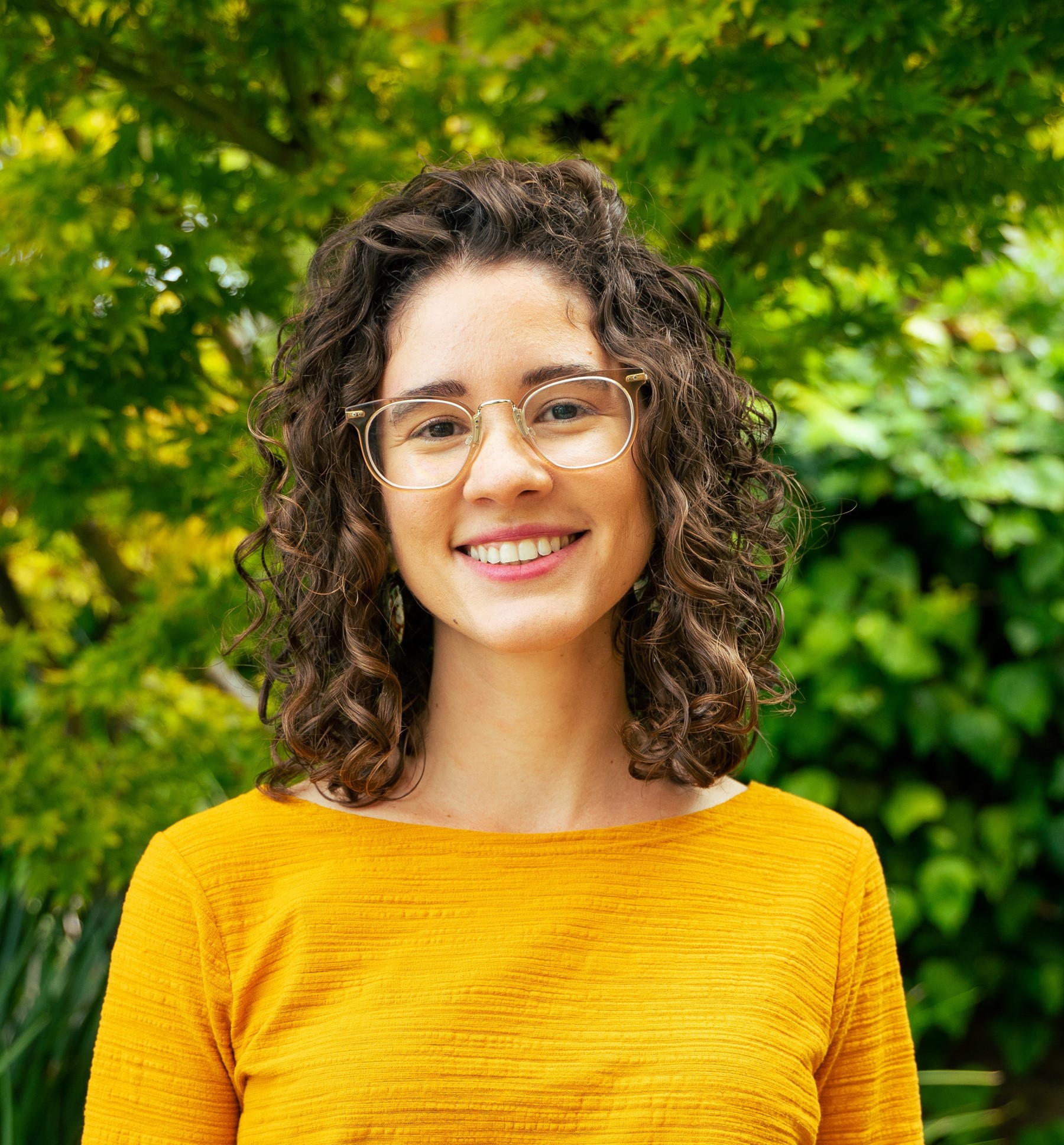 Laurent's headshot, she is a young Latinx woman with shoulder length curly brown hair, a big smile, clear-framed glasses, and a warm yellow top.