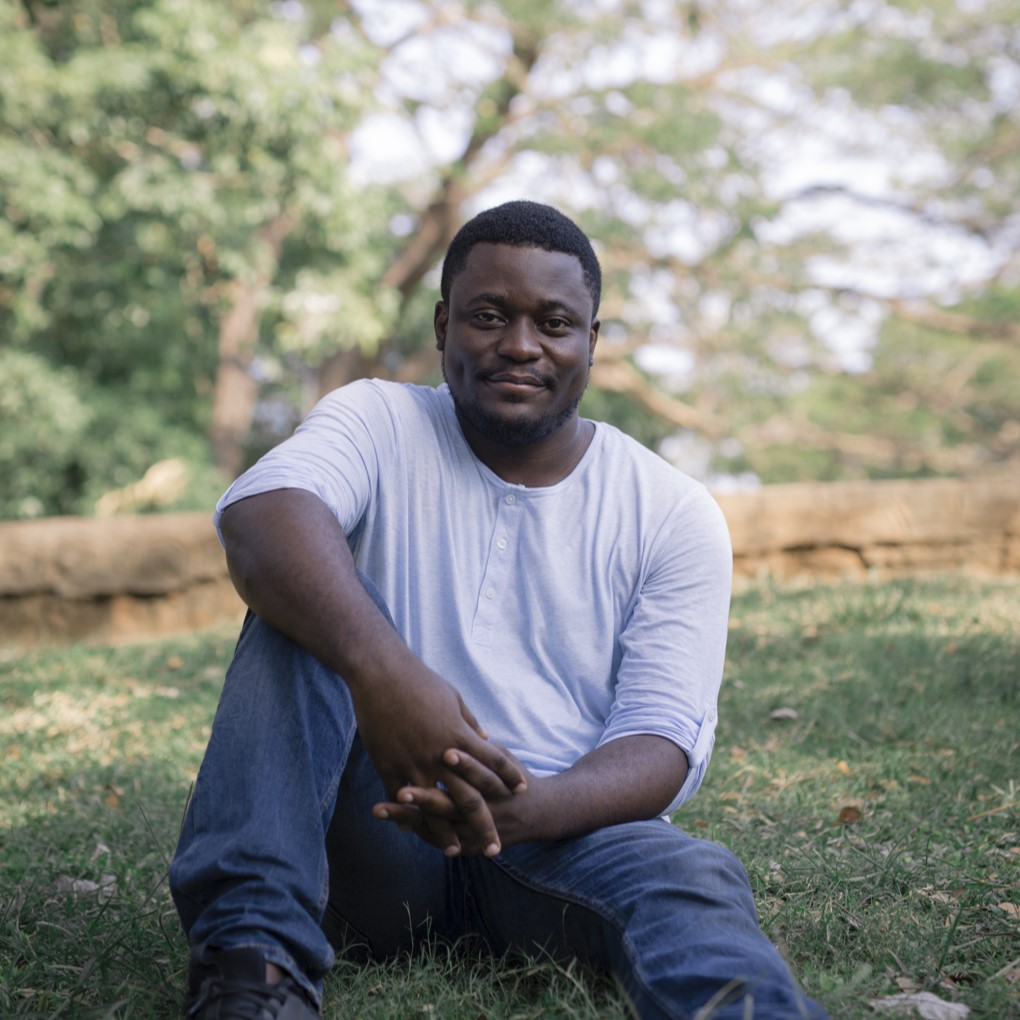 Bayo's headshot; Bayo is a Black man sitting, legs stretched in front of him, in a field of grass. He is gently smiling.