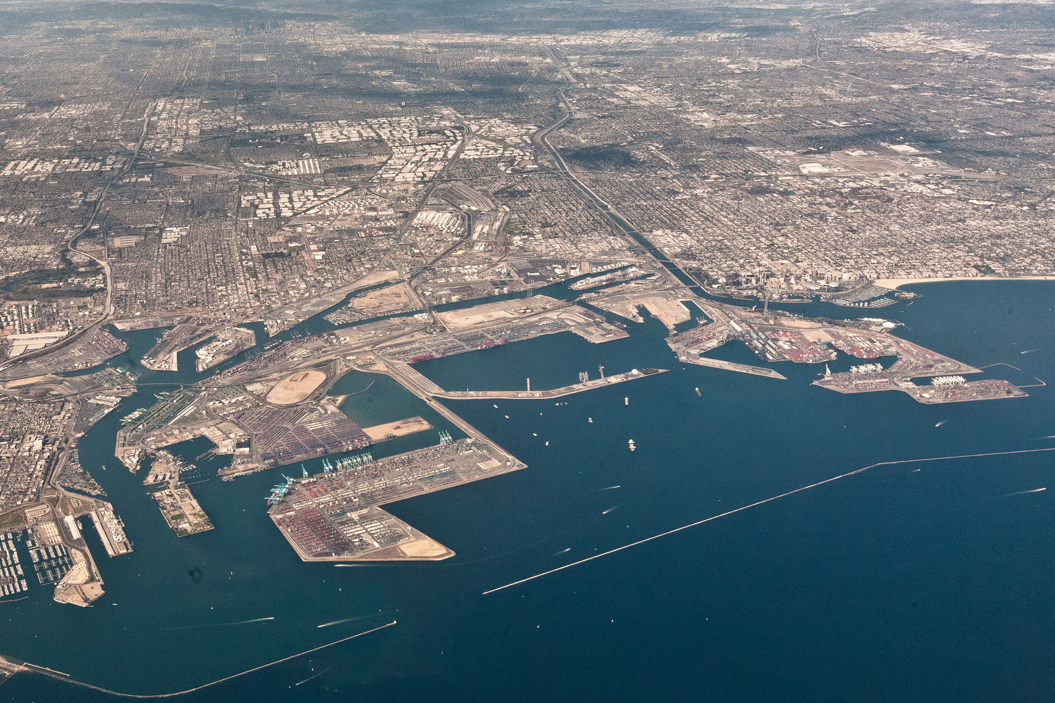 A birds-eye view of the Los Angeles shoreline. Two massive container yards and ports jut out in to the water, lined with many tall cranes. The sprawling urban landscape stretches into the horizon.