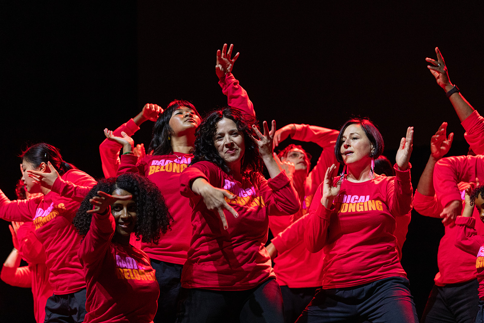 Dancers posing on a stage with a black backdrop wearing red long sleeved shirts that read "Radical Belonging."