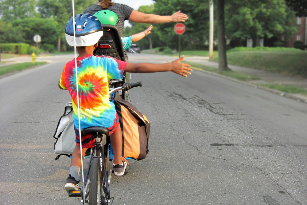 An adult and two children riding on a three-person bike while practicing hand signals for safety (Photo by Mark Stosberg / Unsplash)