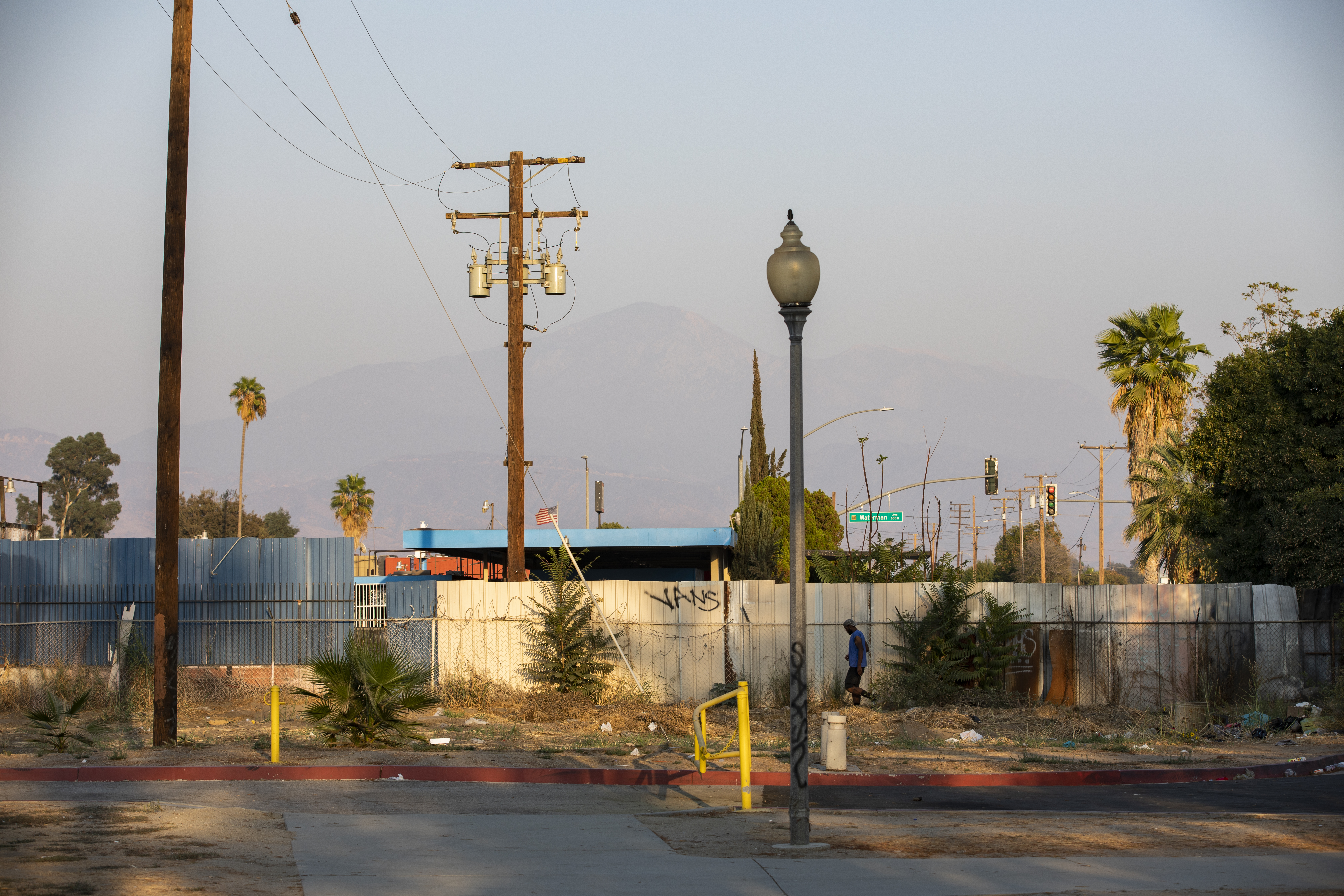 A lone man walks on a tattered, chipped sidewalk abutting a tagged fence. Litter is strewn on the ground and smog hides the mountains in the horizon.