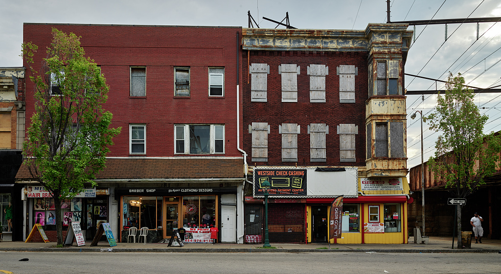 A mixed-use, red brick building in Chester, Pennsylvania. A hair salon, barber shop, and liquor store occupy the ground floor with two stories of apartments sitting on top. Half of the apartments are bordered up, and the building itself appears dilapidated.