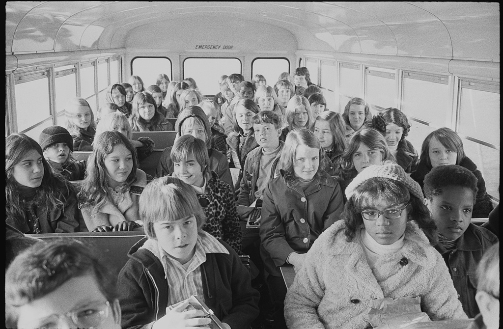 A school bus filled with mostly white children, maybe 40 or so. Some glance at the cameras, some with smirks and others with grimacing faces. Two young black children sit up front, and another black child sits near the back.