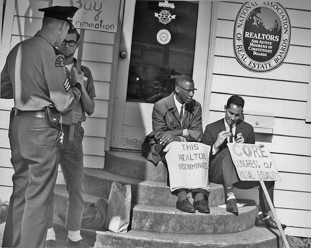 A black and white image of two Black men in suits sitting on the steps of a realtor office with protest placards. A third protestors, a white man, talks with a police officer.