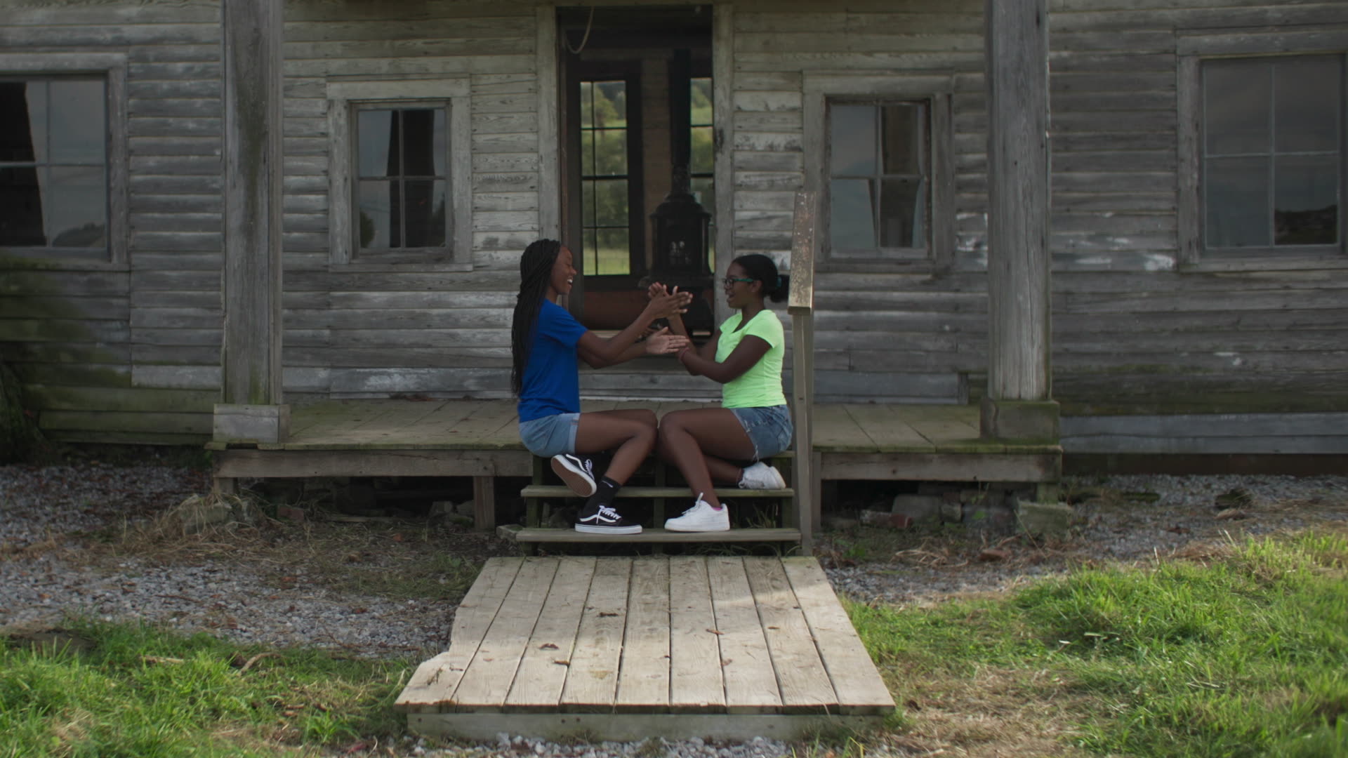 Two young black girls sit profile on a porch playing hand games with each other