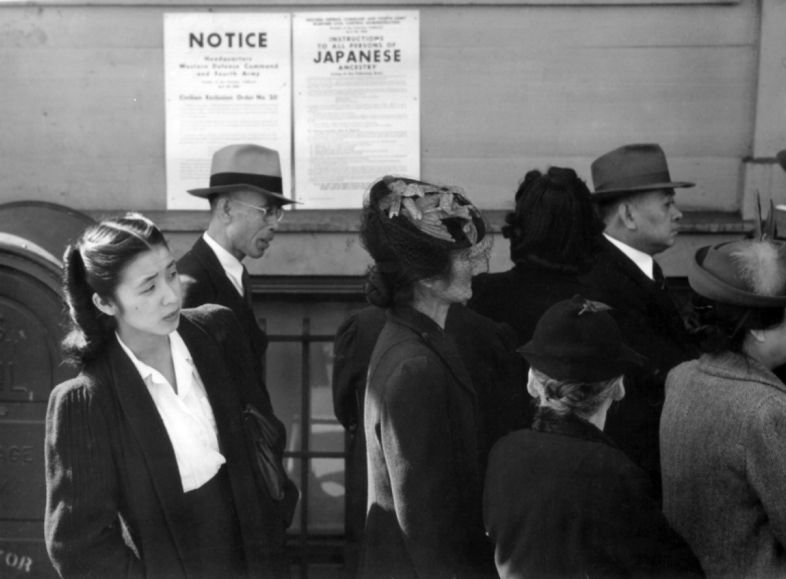 A Black and white image of Japanese Americans standing in line. Behind them, a poster is pinned against the wall stating that people of Japanese ancestry will be interned. A woman peeks her head out to look to the front of the line,. 