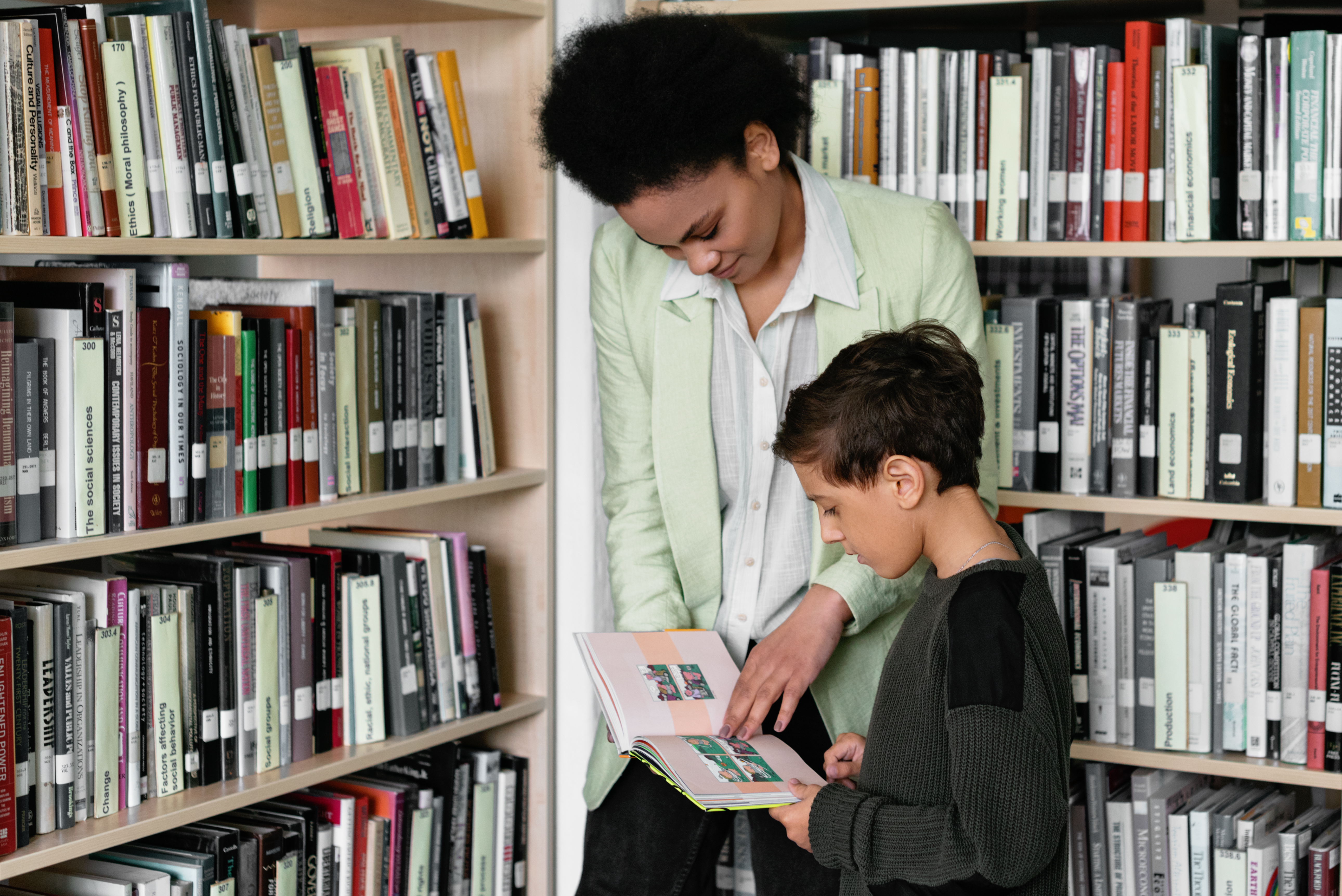 A child and a young woman read through a picture book in a library. They're surrounded by books.