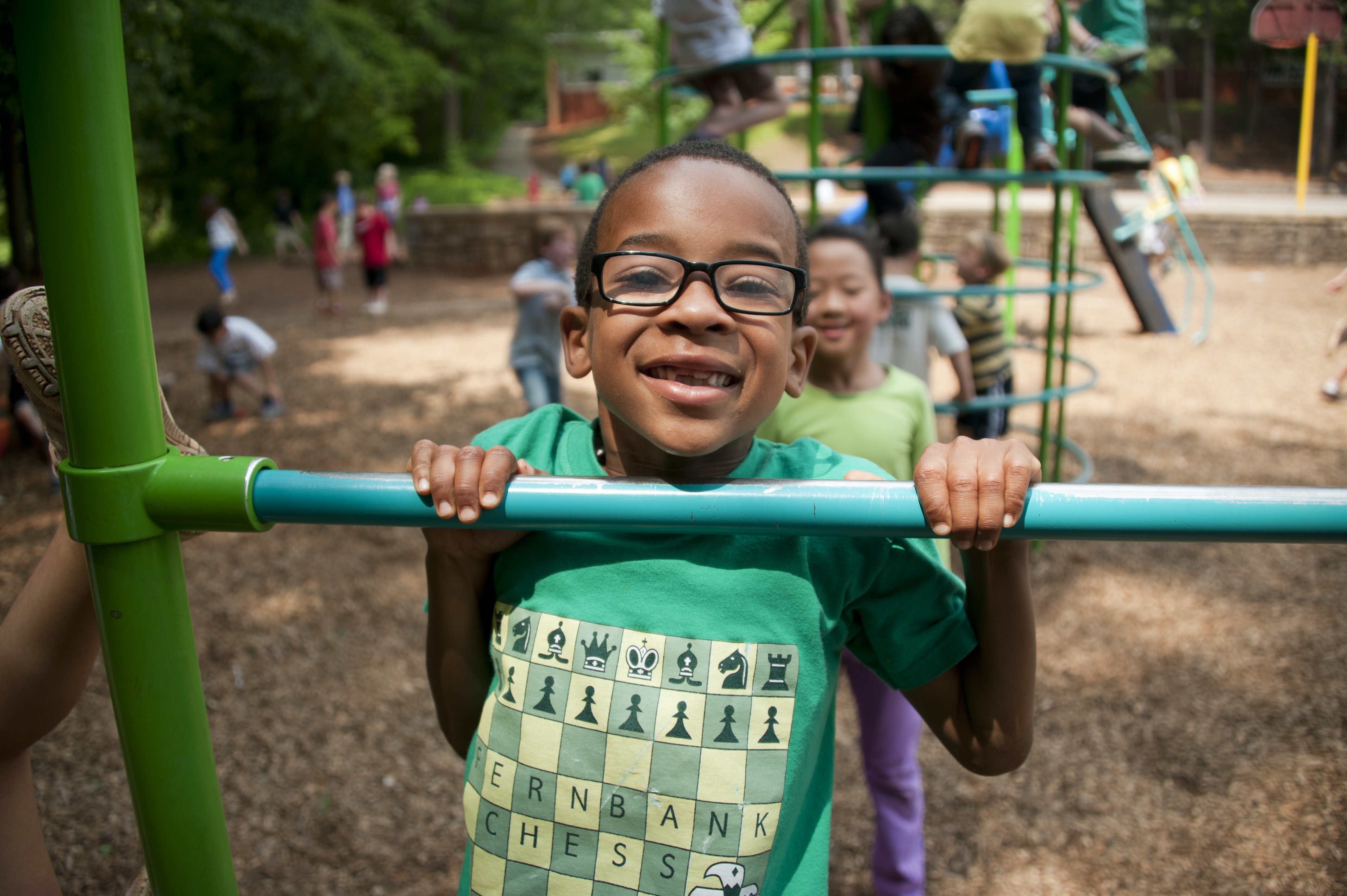 A young Black boy wearing glasses and a green shirt smiles a big toothy grin as he pulls himself up a bar in a playground.