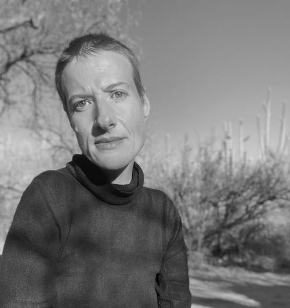 A black-and-white photograph off Sunaura Taylor, a slender woman wearing short cropped hair and a dark turtleneck. She is outside on a bright day, and her head is slightly cocked looking at the camera.