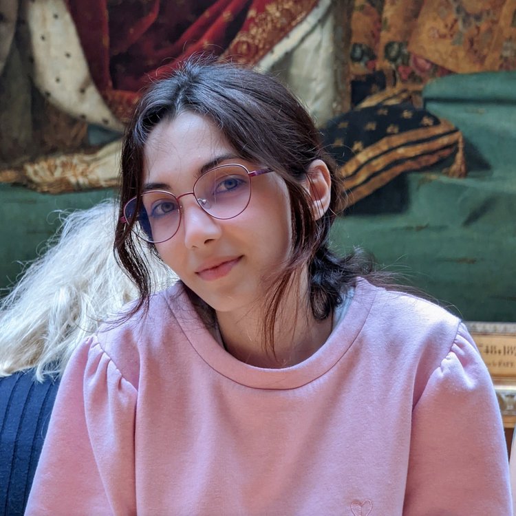 Lara Habboub’s headshot, a young Syrian Palestinian woman wearing wireframe glasses and a pink sweater. She looks at the camera with a small smile.
