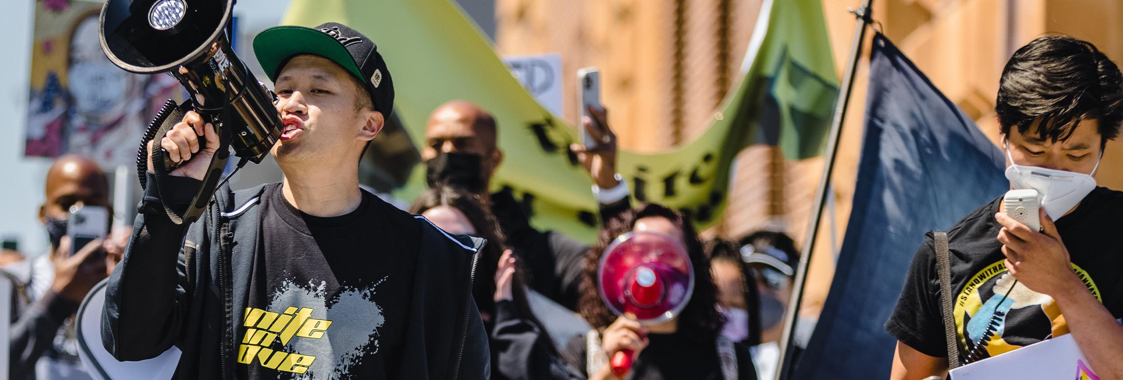An Asian man chants into a bullhorn at the head of a demonstration against anti-Asian hate. A crowd holding banners follow him.