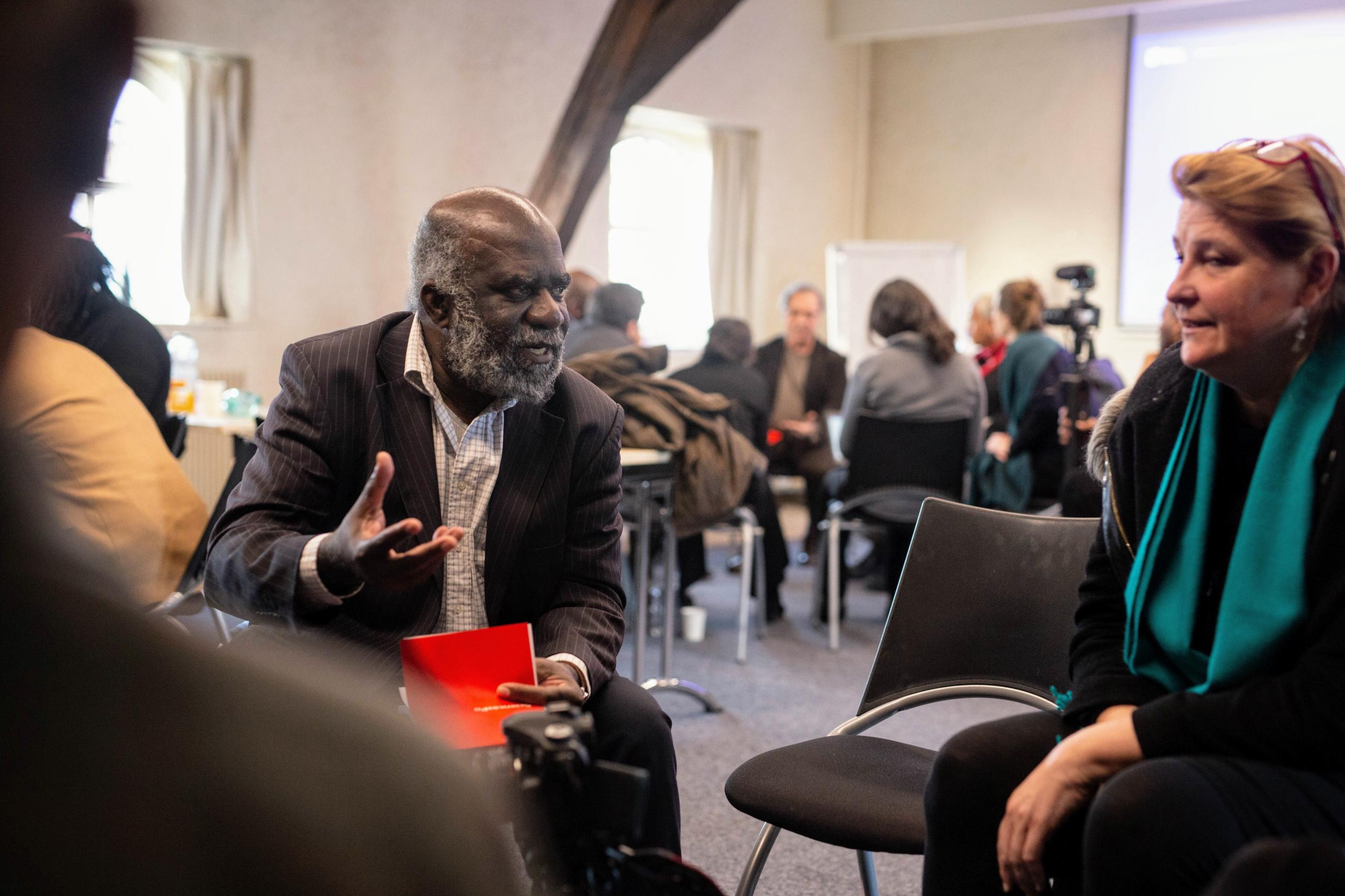 Two people talking at the Othering & Belonging Paris symposium in January 2020. One is a Black man wearing a suit, the other is a white woman with a green scarf and black shirt.