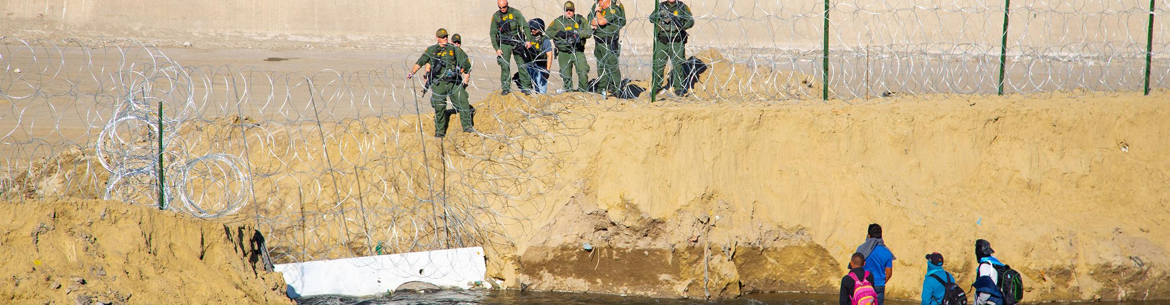 A group of migrants looks up at a barbed-wire fence and border police