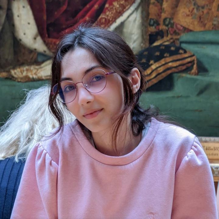 Lara Habboub’s headshot, a young Syrian Palestinian woman wearing wireframe glasses and a pink sweater. She looks at the camera with a small smile. 